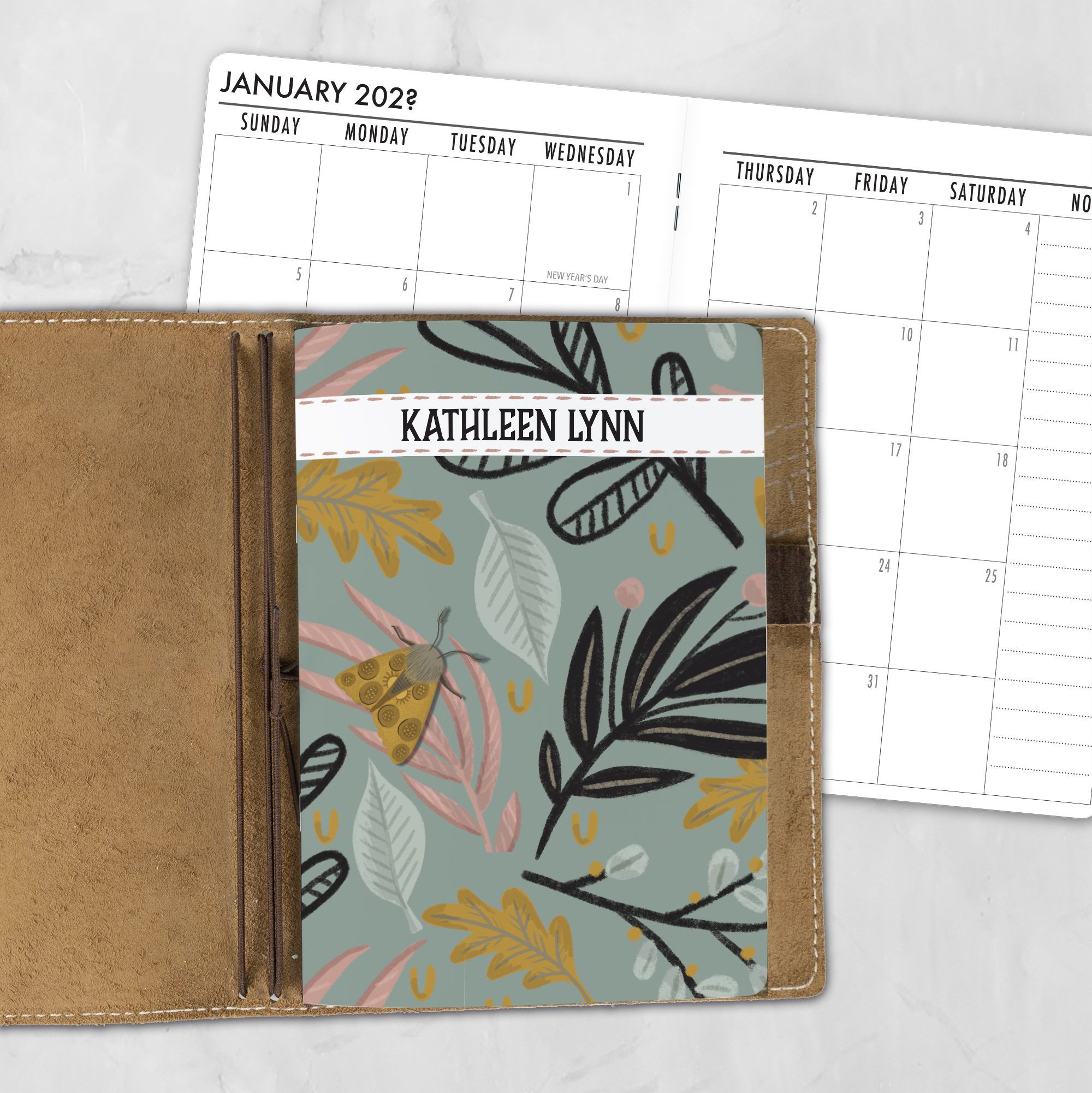 A5 Moth Fern personalized traveler's notebook refill showing the cover design inside a leather traveler's notebook cover, with a monthly calendar spread visible behind.