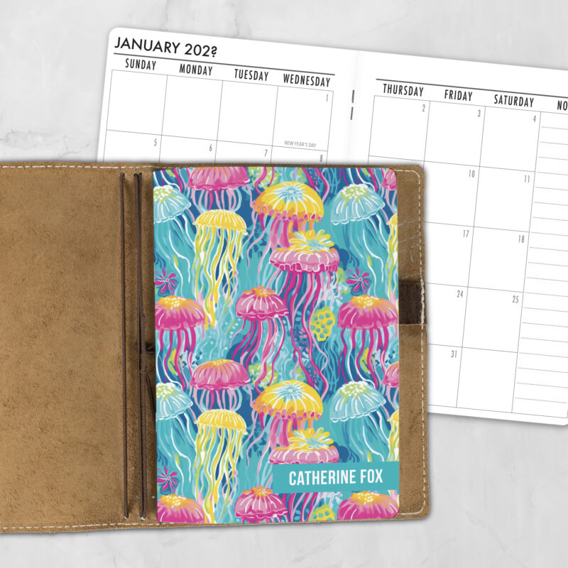 A5 Neon Jellyfish personalized traveler's notebook refill showing the cover design inside a leather traveler's notebook cover, with a monthly calendar spread visible behind.