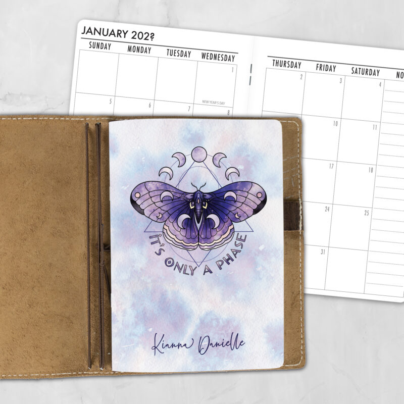 A5 Only a Phase personalized traveler's notebook refill showing the cover design inside a leather traveler's notebook cover, with a monthly calendar spread visible behind.