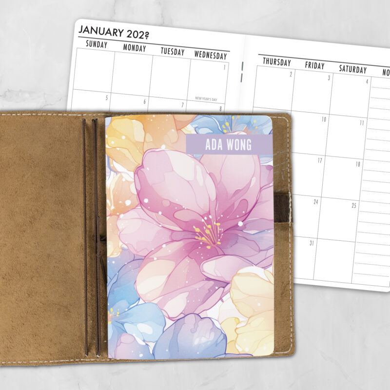 A5 Spring Petals personalized traveler's notebook refill showing the cover design inside a leather traveler's notebook cover, with a monthly calendar spread visible behind.