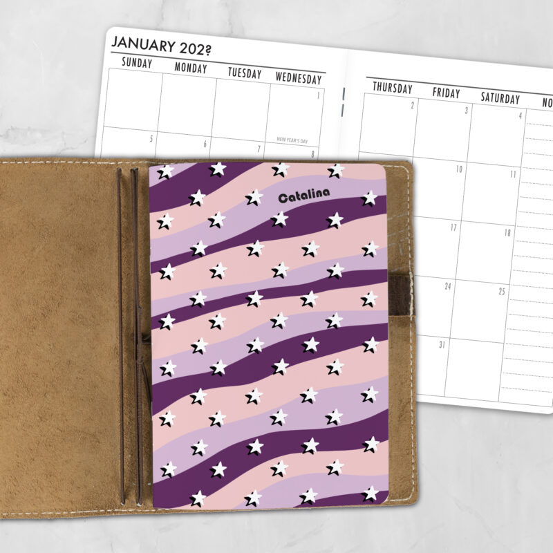 A5 Stardust personalized traveler's notebook refill showing the cover design inside a leather traveler's notebook cover, with a monthly calendar spread visible behind.