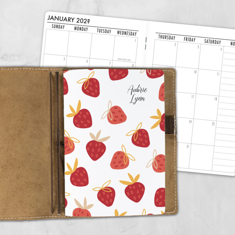 A5 Strawberry Crush personalized traveler's notebook refill showing the cover design inside a leather traveler's notebook cover, with a monthly calendar spread visible behind.