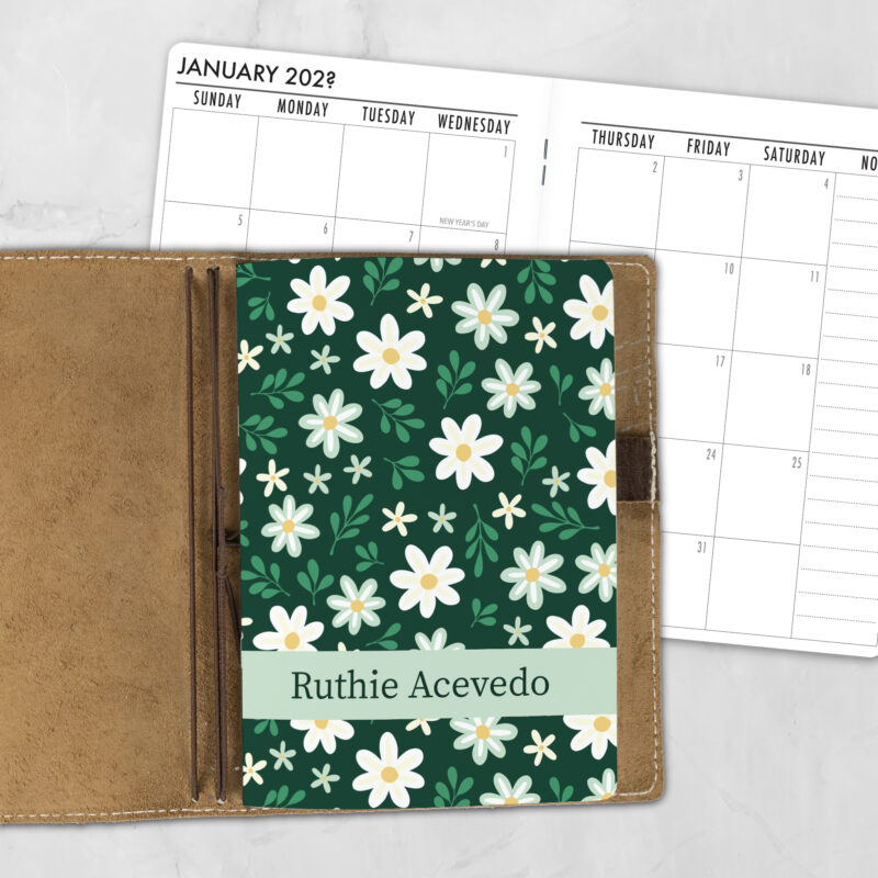A5 Sunday Daisies personalized traveler's notebook refill showing the cover design inside a leather traveler's notebook cover, with a monthly calendar spread visible behind.