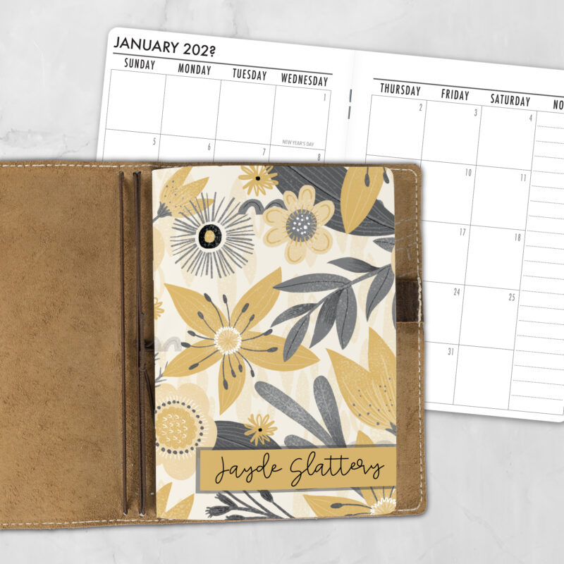A5 Sunglow personalized traveler's notebook refill showing the cover design inside a leather traveler's notebook cover, with a monthly calendar spread visible behind.