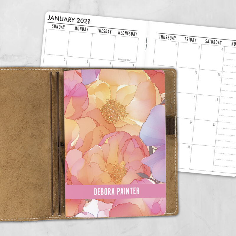 A5 Sunset Blooms personalized traveler's notebook refill showing the cover design inside a leather traveler's notebook cover, with a monthly calendar spread visible behind.