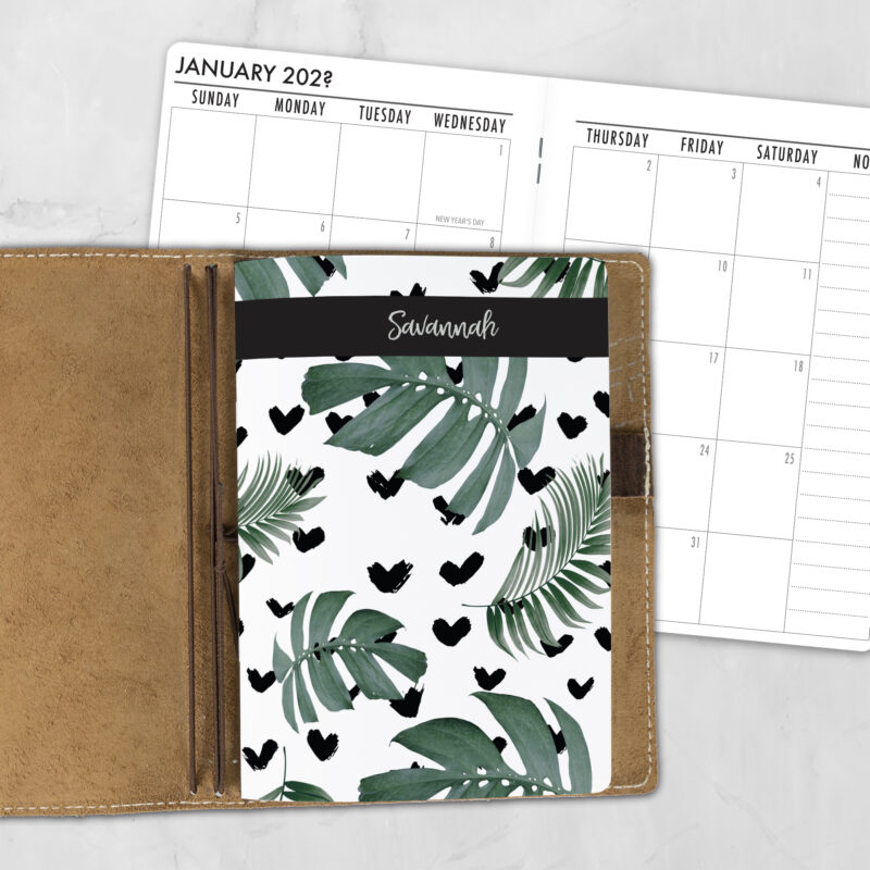 A5 Tropical Love personalized traveler's notebook refill showing the cover design inside a leather traveler's notebook cover, with a monthly calendar spread visible behind.