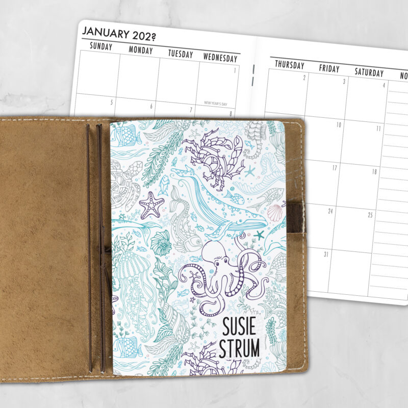 A5 Underwater Seaglass personalized traveler's notebook refill showing the cover design inside a leather traveler's notebook cover, with a monthly calendar spread visible behind.