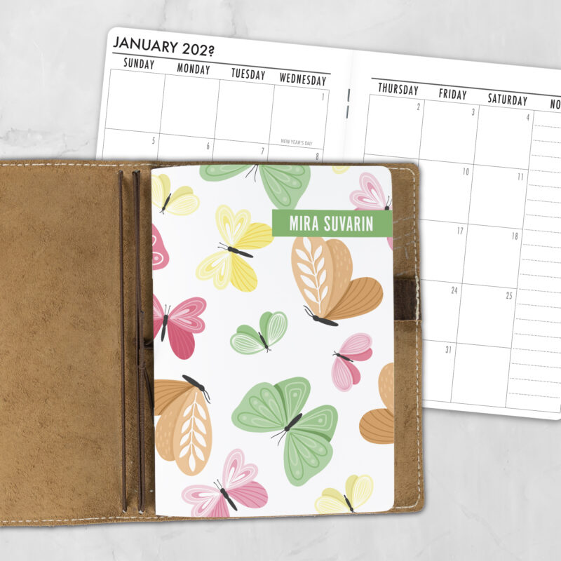A5 Whimsical Wings personalized traveler's notebook refill showing the cover design inside a leather traveler's notebook cover, with a monthly calendar spread visible behind.