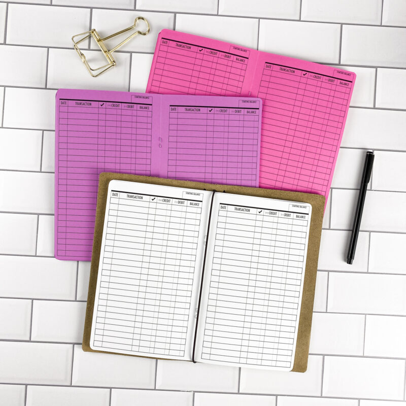 Checkbook Register traveler's notebook refill by CityGirl Planners, showing checkbook register pages with columns for transactions, deposits, and balances. Three booklets displayed in different paper color options inside a traveler's notebook cover.