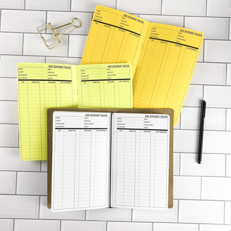 Debt Repayment traveler's notebook refill by CityGirl Planners, showing debt repayment tracker pages for managing payoff progress. Three booklets displayed in different paper color options inside a traveler's notebook cover.