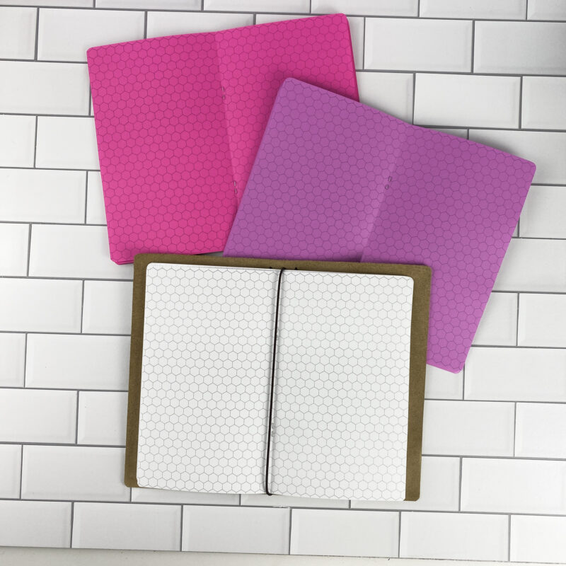 Hex Grid traveler's notebook refill by CityGirl Planners, showing hexagonal grid pages for creative note taking and game mapping. Three booklets displayed in different paper color options inside a traveler's notebook cover.