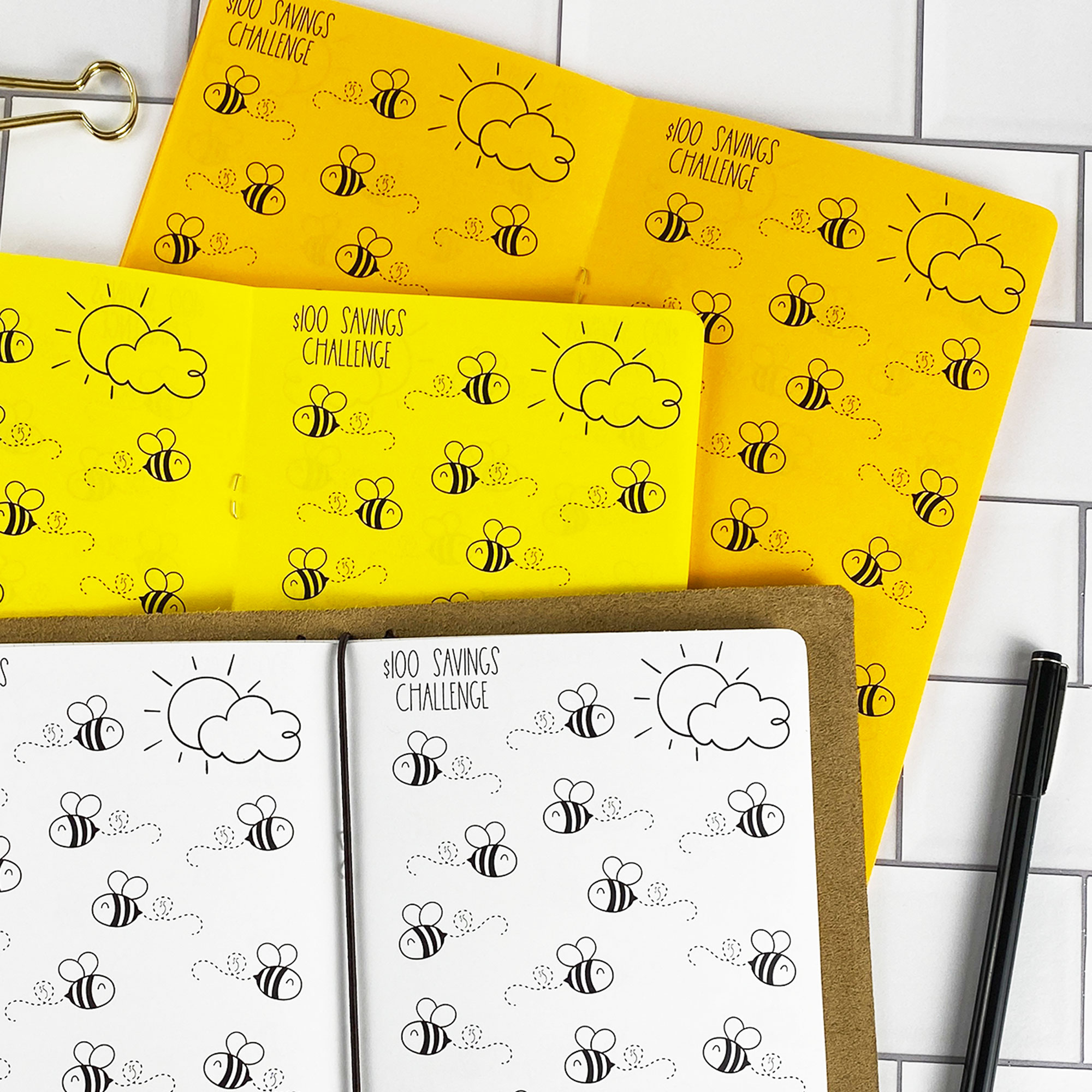Detail view of the Savings Challenge Buzzin Bees traveler's notebook refill showing $100 savings challenge pages with cute bee illustrations to color in as you save, displayed with multiple paper color options.