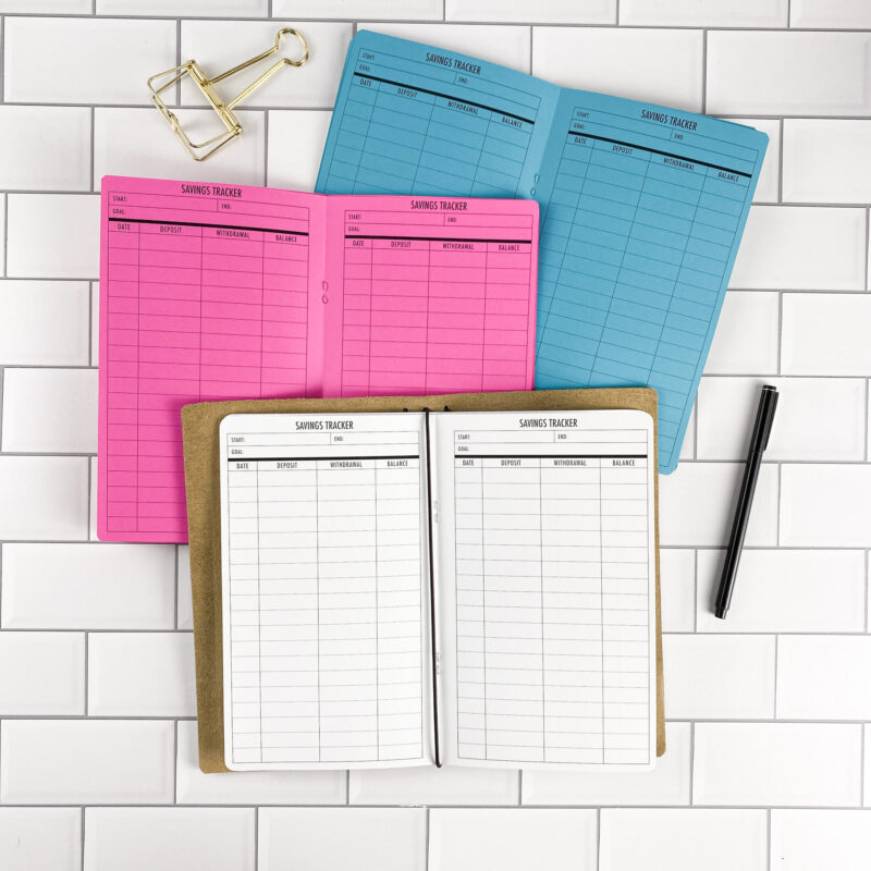 Savings Tracker traveler's notebook refill by CityGirl Planners, showing savings tracker pages with columns for deposits, withdrawals, and balance. Three booklets displayed in different paper color options inside a traveler's notebook cover.