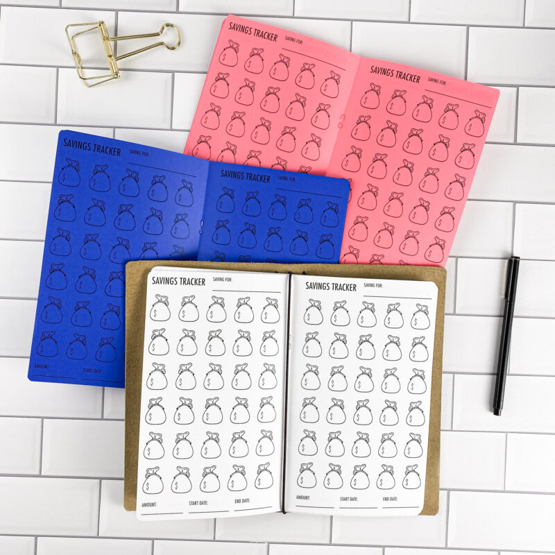 Savings Tracker Money Bags traveler's notebook refill by CityGirl Planners, showing savings tracker pages with a money bag illustration design. Three booklets displayed in different paper color options inside a traveler's notebook cover.