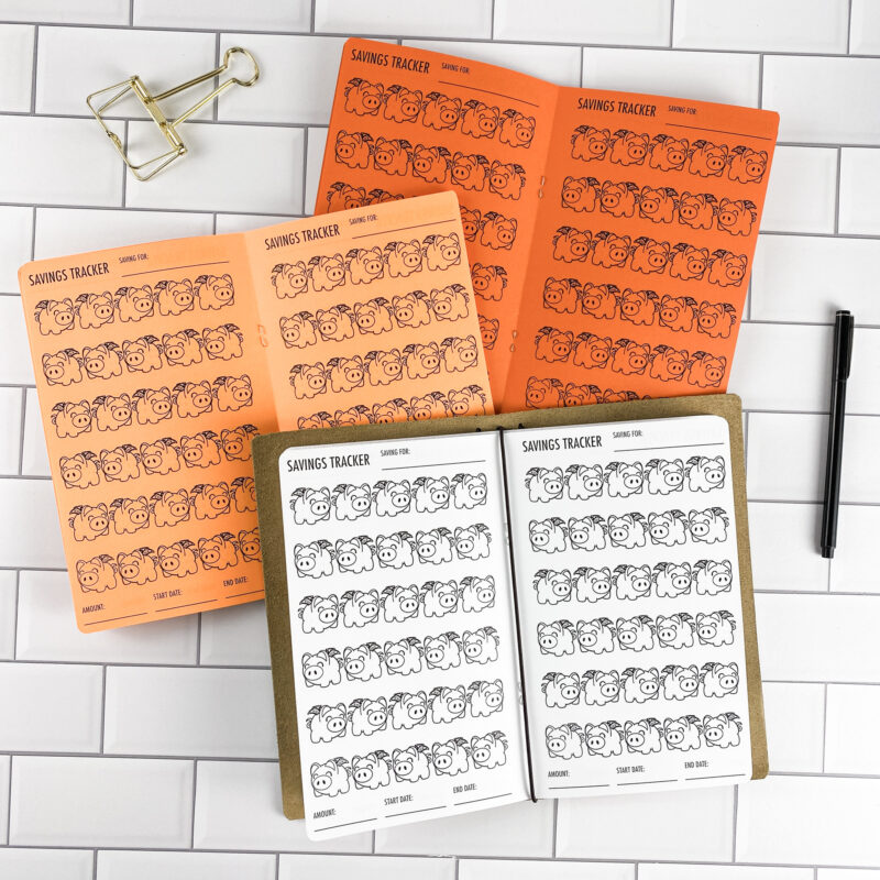 Savings Tracker Piggy Banks traveler's notebook refill opened to show the savings tracker pages with a piggy bank illustration design in bright white paper, saddle-stitched booklet format.